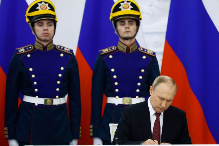 Two soldiers in ceremonial uniform standing to attention with Russian flags behind them. Putin is in front signing the 'annexation' of four Ukrainian territories partly occupied by Russia.