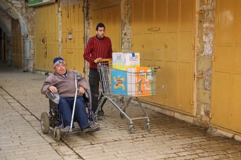 Qafisheh rolls along next to his son Osama as they bring a shopping cart of restock past the checkpoints to the shop