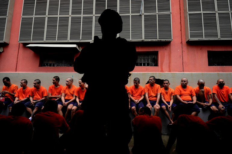 A soldier stands in shadow in front of a row of orange-clad prisoners.